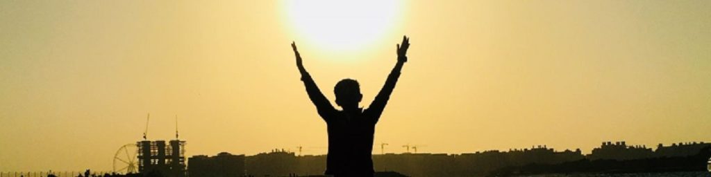 A child raising both hands in the air, with the sun visible between them
